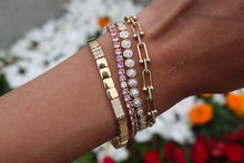 Load image into Gallery viewer, Close-up of a wrist wearing multiple gold, diamond, tennis, and pink sapphire bracelets with a blurred floral background
